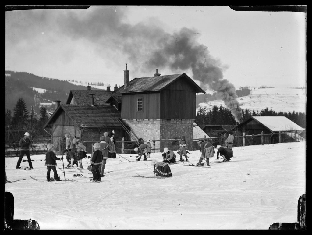 Czarnobiałe zdjęcie. Grupa osiemnastu osób, w tym trzy osoby dorosłe i piętnaścioro dzieci. Dzieci zapinają narty. Na drugim planie zabudowania dworca kolejowego i dym z lokomotywy. Tłem są zbocza Pasma Gubałowskiego.