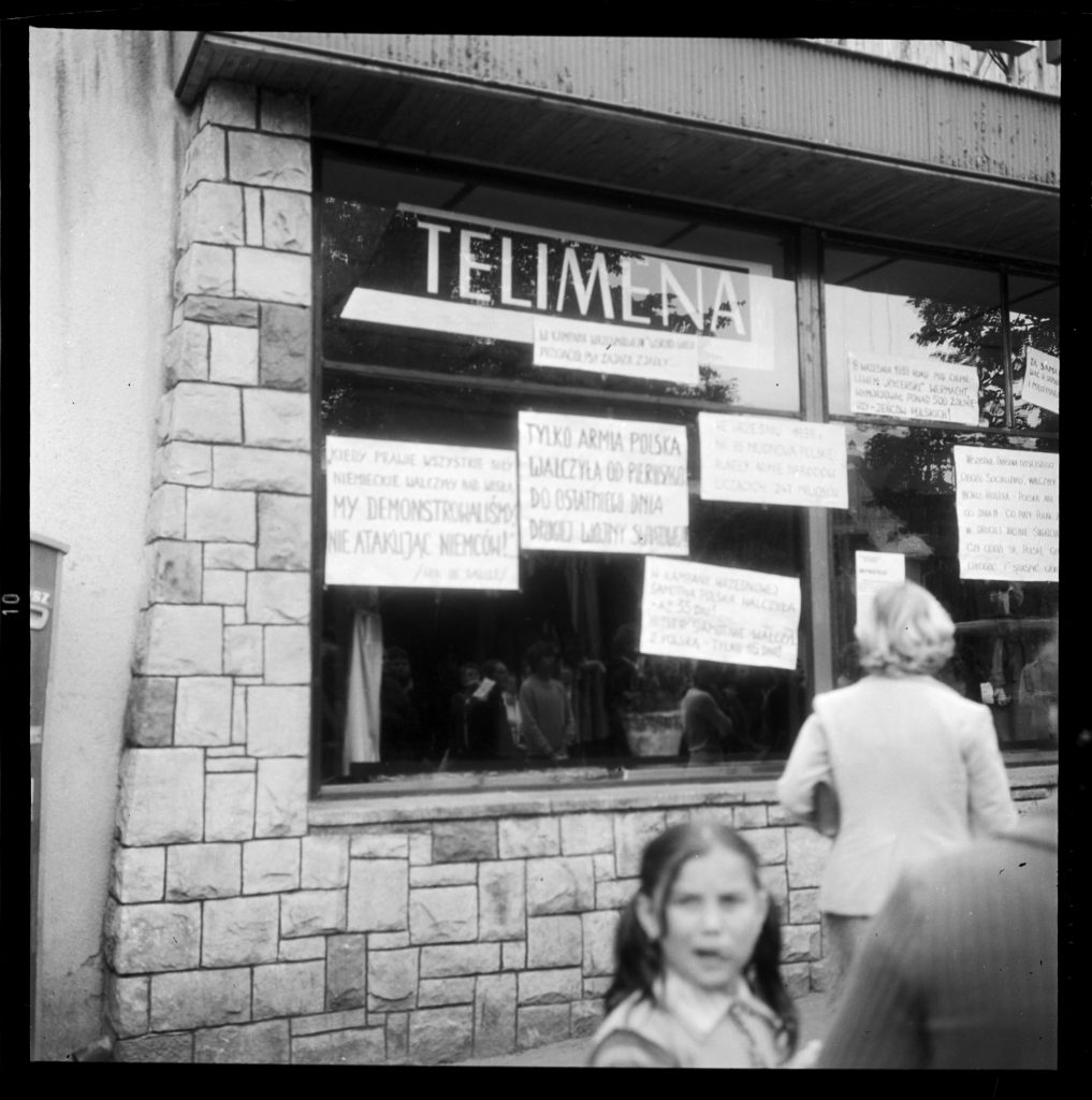 Czarno - biała fotografia - widoczni przechodnie i afisze w witrynie sklepu Telimena