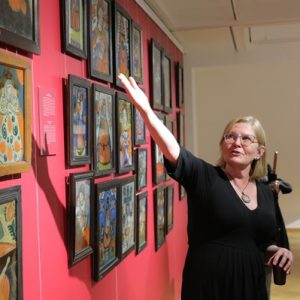 kobieta w czarnej sukience wskazuje ręką obrazy malowane na szkle