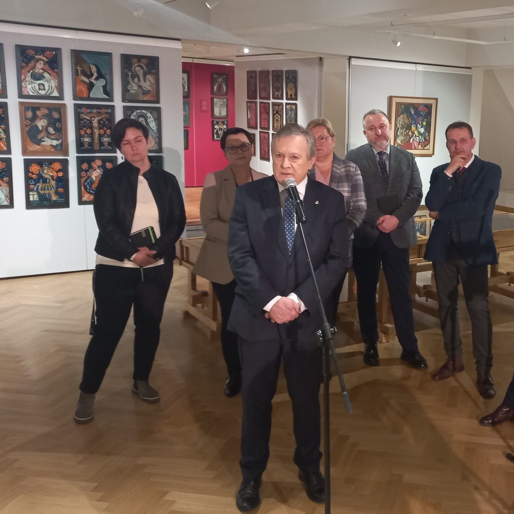 mężczyzna stoi przed mikrofonem, za nim kilku mężczyzn i kobiet wokół ekspozycja obrazów malowanych na szkle