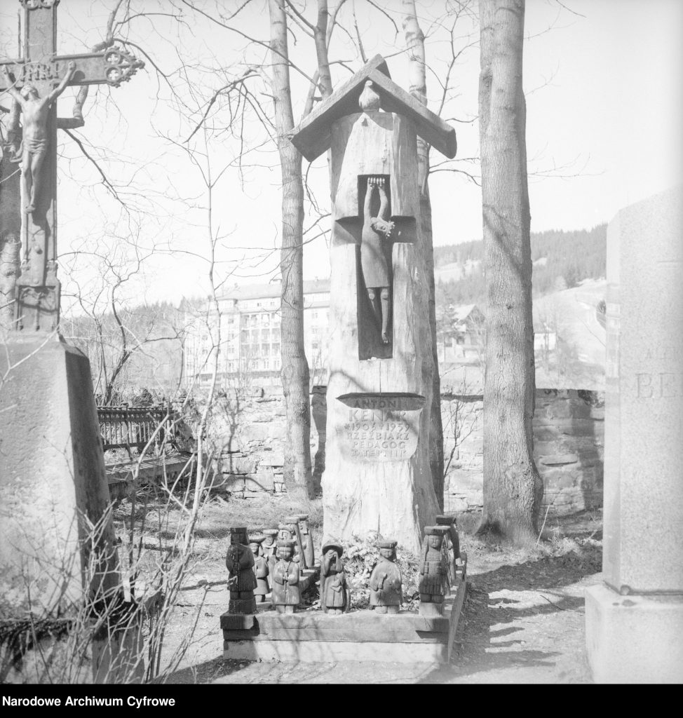 Czarno-białe zdjęcie przedstawia fragment cmentarza na Peksowym Brzyzku w Zakopanem. W centrum widoczny drewniany nagrobek Antoniego Kenara