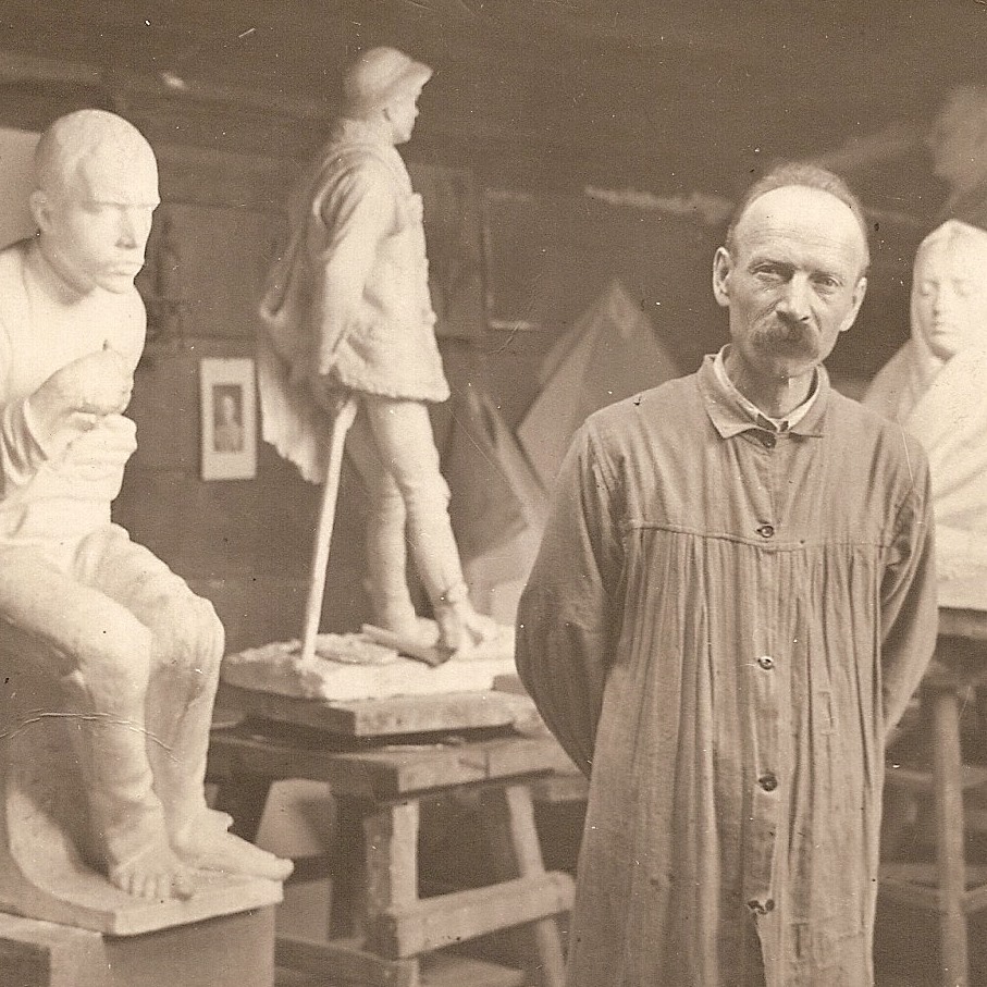 czarno białe zdjęcie. Mężczyzna z założonymi do tyłu rękami stoi wśród rzeźb z drewna
