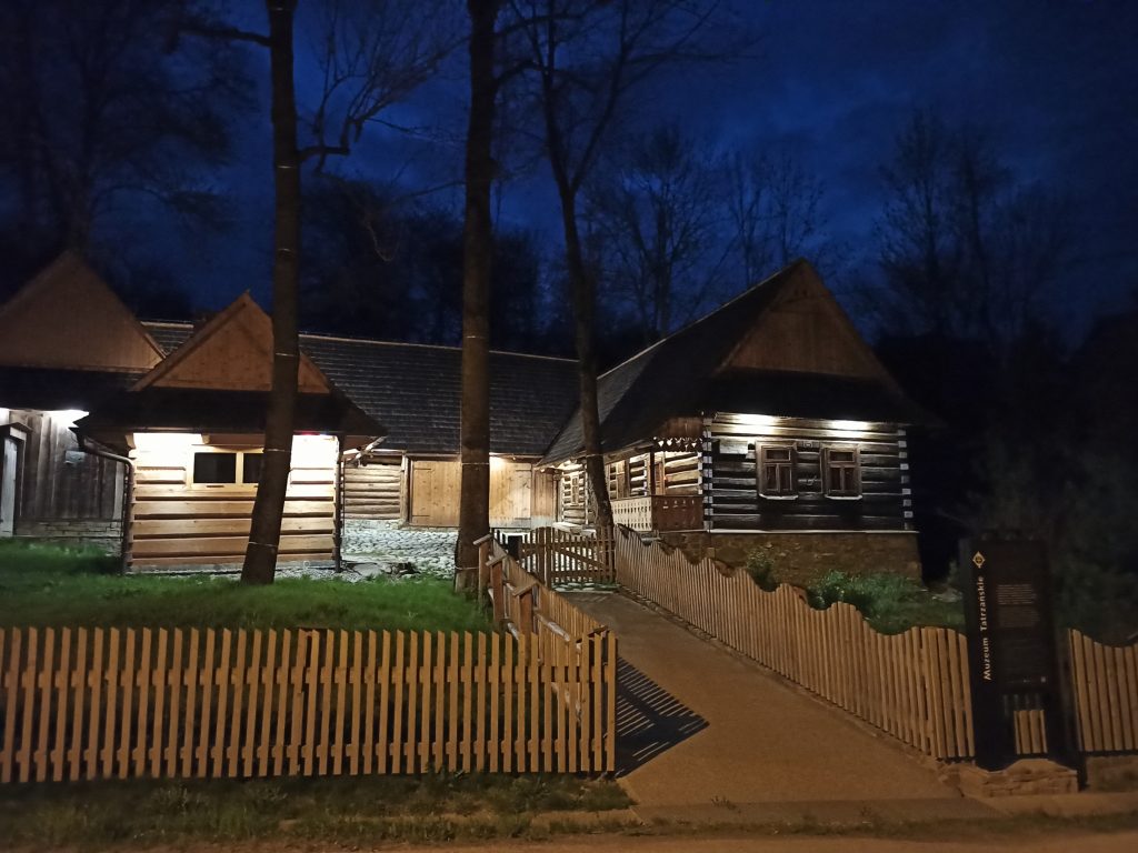 drewniane budynki otoczone drewnianym płotem, podświetlone w nocy
