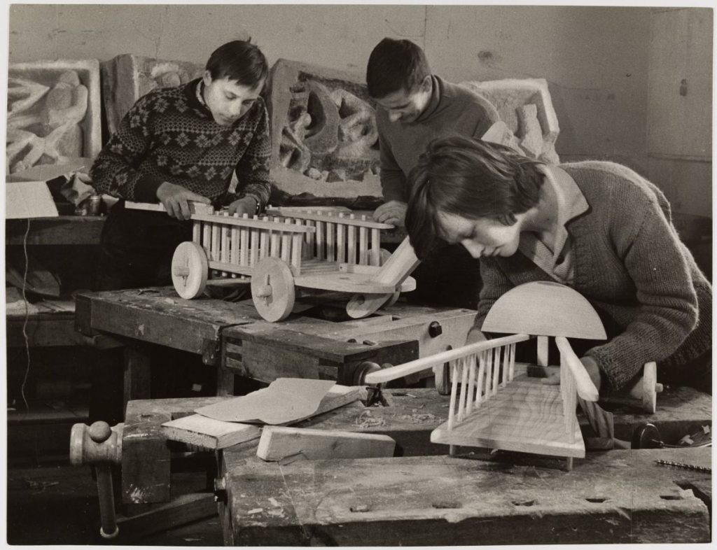 Czarno-białe zdjęcie przedstawia trzech chłopców w warsztacie tworzących drewniane pojazdy