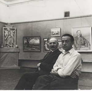 czarno białe zdjęcie: dwaj mężczyźni siedzą w sali wystawowej, za nimi obrazy i rzeźby