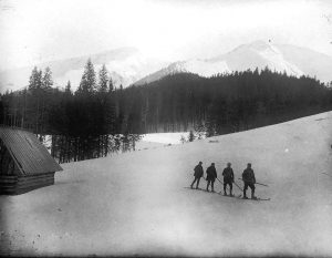 Czterej narciarze na hali koło szałasu na tle lasu - czarno-biała fotografia