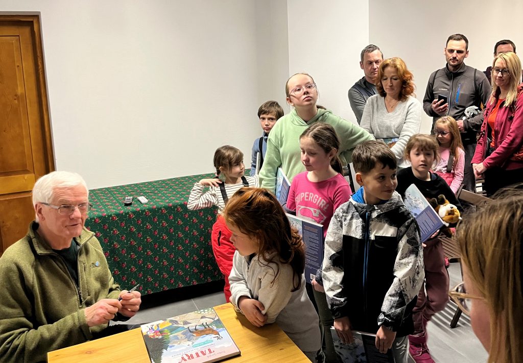 Paweł Skawiński podpisuje książkę, obok grupa dzieci z rodzicami czekający w kolejce