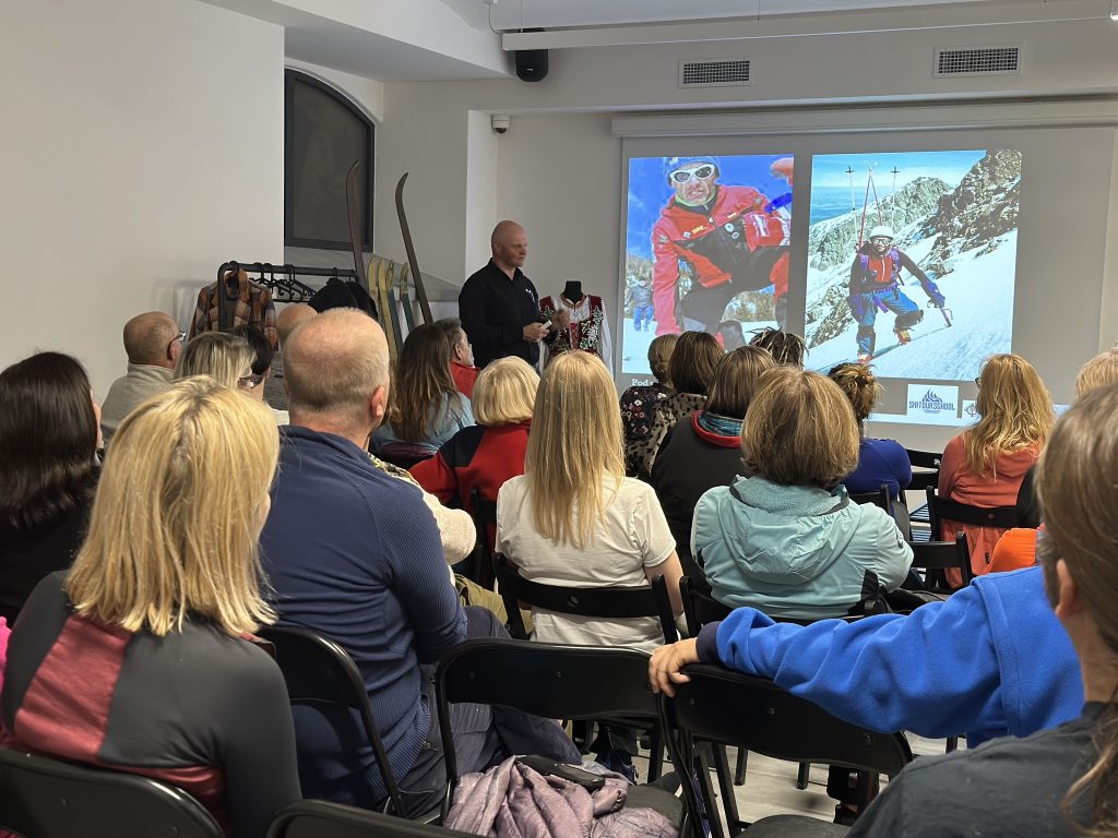 Wojciech Szatkowski opowiadza licznie zebranym gościom siedzącym na krzesłach w sali muzeum. Opowieść o narciarstwie ilustrowana jest slajdami z wypraw, na których widać narciarzy. Na pierwszym planie kilkadziesiat osób siedzacych i słuchających, w tle prowadzący pokazujący slajdy.