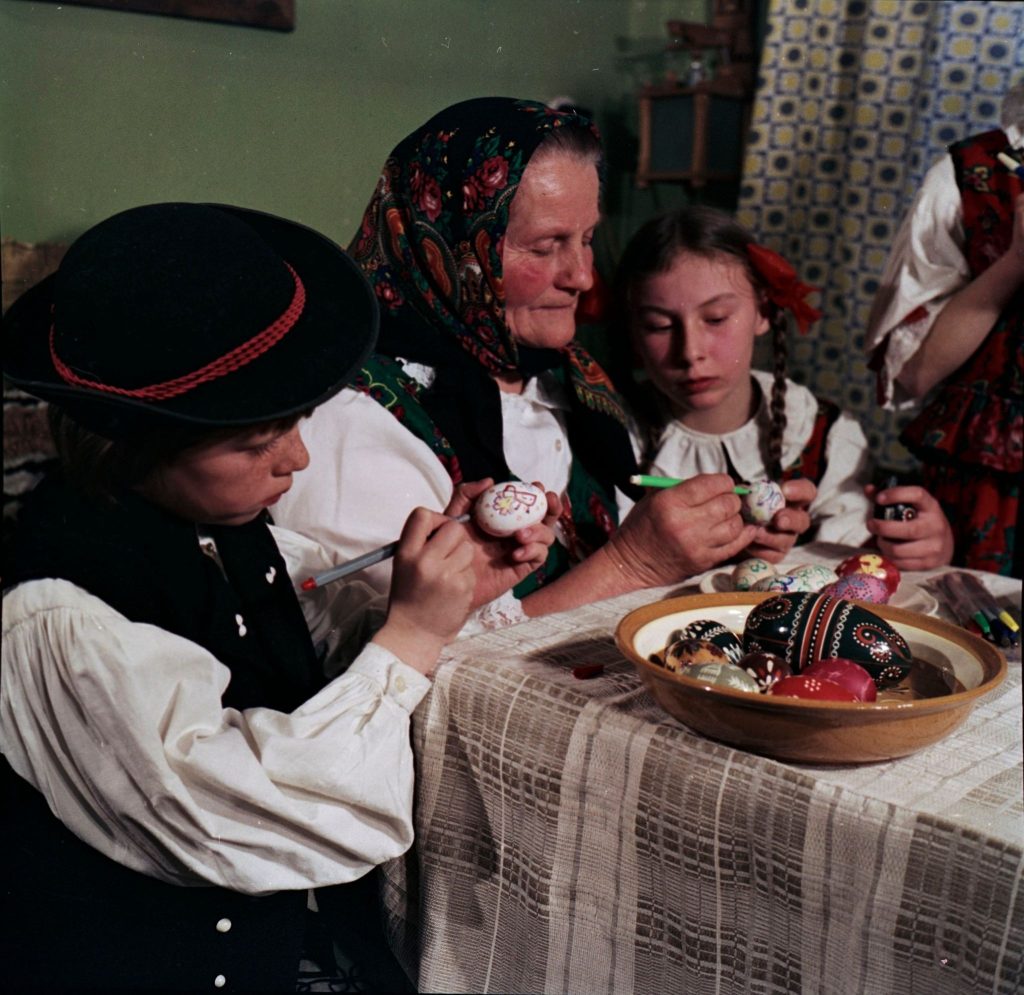 Wielkanoc Tradycyjne zdobienie pisanek na Orawie. Fotografia przedstawia starszą Panią i dwójkę dzieci w regonalnych strojach siedzących przy stole i ozdabiających pisanki. fot. Władysław Werner (3)
