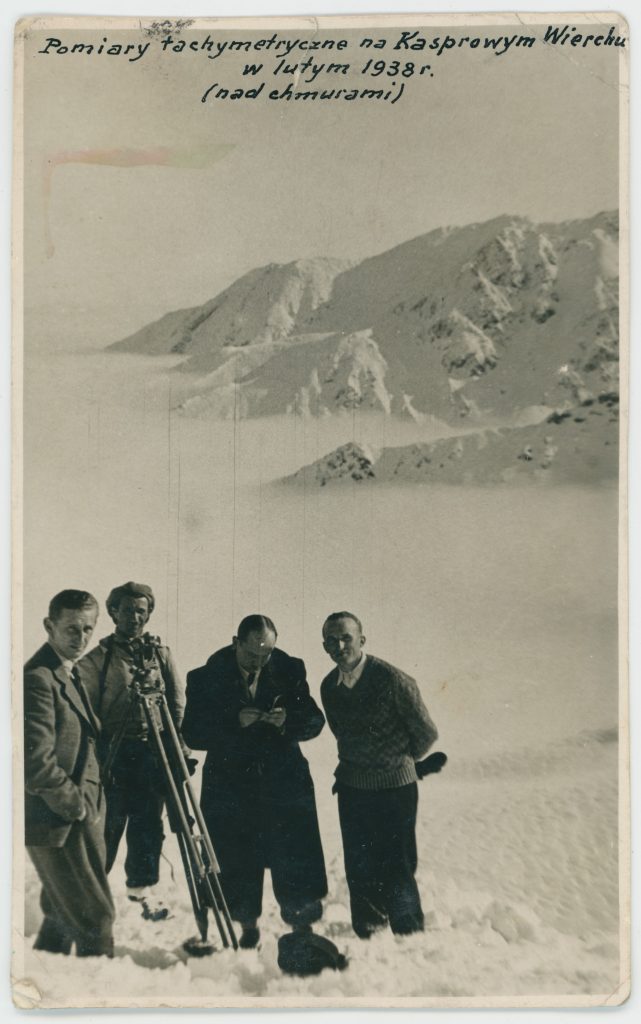 Czarno-białe zdjęcie czterech mężczyzn na tle zaśnieżonych gór podczas budowy kolejki na Kasprowy Wierch, nad nimi napis "pomiary techniczne na Kasprowym Wierchu w lutym 1938 (nad chmurami)"