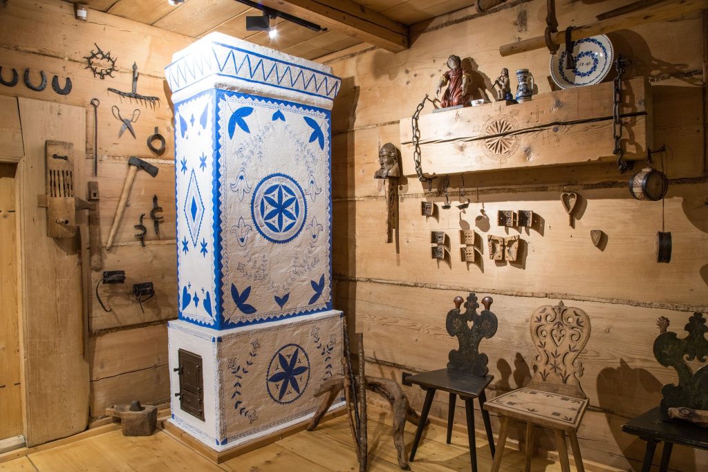 Wnętrze góralskiej chałupy, drewniane meble krzesła, skrzynia i półki oraz bielony piec i eksponaty wiszące na ścianach