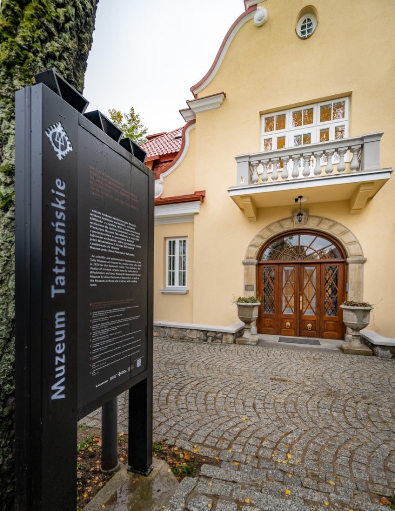 Budynek Galerii na Kozińcu \ Na pierwszym planie czarny znak Muzeum Tatrzańskiego. Na drugim planie budynek galerii z żółtą fasadą