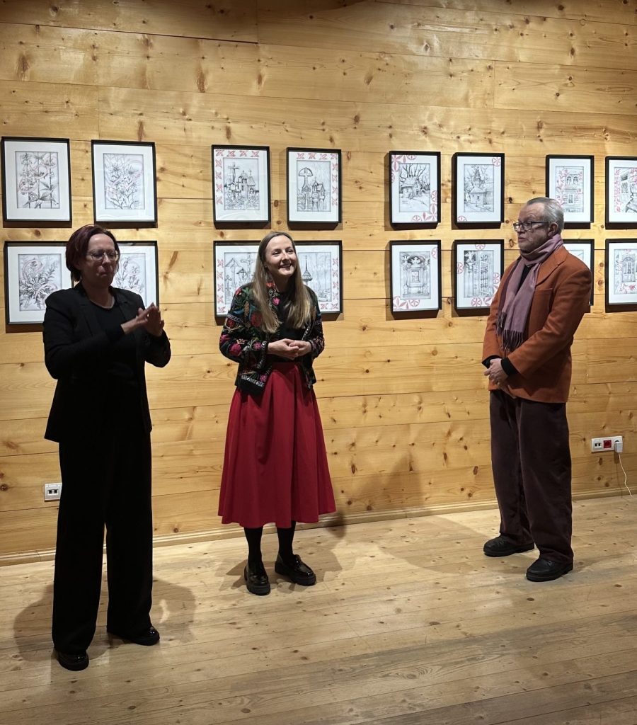 Na zdjęciu stoją trzy osoby - kuratorzy wystawy: Marek Krzysztof Lasecki i dr Emilia Pomiankiewicz-Wagner oraz tłumaczka języka migowego na tle rysunków prezentowanych na wystawie „SpotkajMY się w MT”.