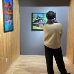 Na zdjęciu osoba ogląda obrazy prezentowane na wystawie „SpotkajMY się w MT”. Na ścianach widoczne są kolorowe obrazy przedstawiające budynki.