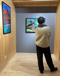 Na zdjęciu osoba ogląda obrazy prezentowane na wystawie „SpotkajMY się w MT”. Na ścianach widoczne są kolorowe obrazy przedstawiające budynki.