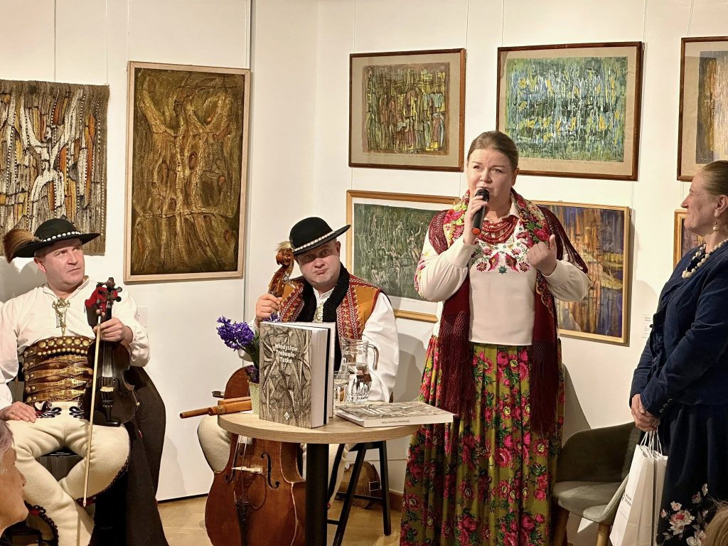 Sala wystawy ,,Władysław Trebunia-Tutka. Tutaj, poza czas”. W tle obrazy Władysława Trebuni-Tutki. Uczestnicy wystawy to: dwóch synów Władysława Trebuni-Tutki, ubranych w tradycyjne stroje góralskie, córka mówiąca do mikrofonu również w tradycyjnym stroju oraz stojąca obok niej kuratorka wystawy - Helena Pitoń.